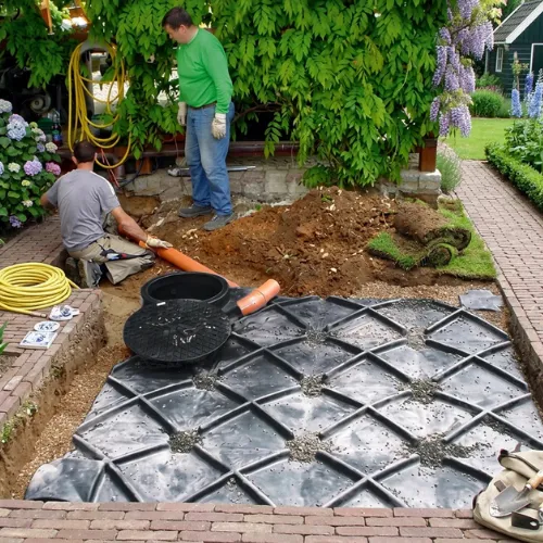 Installatie van een grote regenwaterput van 5000 liter in een uitgegraven put in de tuin waarbij werkmannen pvc afvoerbuizen aansluiten.
