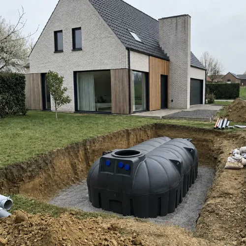 Een grote regenwaterput van 7500 liter in kunststof geplaatst in een open, uitgegraven put op een bed van stabilisé met de blauwe aansluitpunten gericht naar de woning.